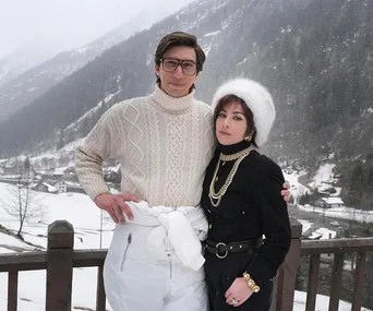 A couple stands in snowy mountains; man in glasses and sweater, woman in black with fur hat and gold jewelry.