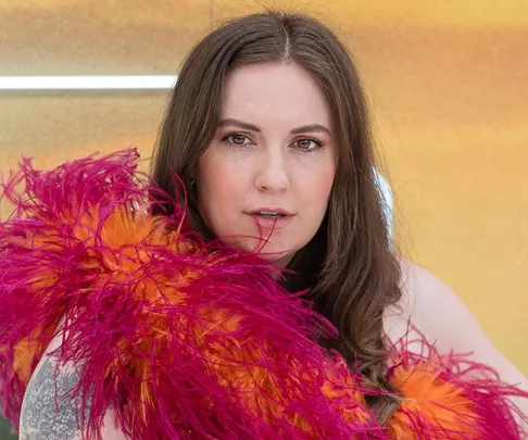 Woman with long brown hair, wearing a vibrant feathered scarf against a warm, blurred background.