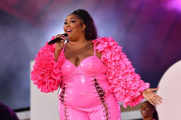 A woman in a bright pink, ruffled outfit sings on stage, holding a microphone, with a background of colorful lights.