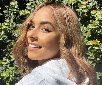 Smiling woman with wavy hair in sunlight, standing in front of green leafy background.