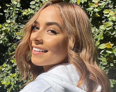 Smiling woman with wavy hair in sunlight, standing in front of green leafy background.