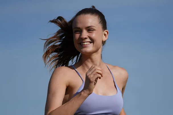 Person smiling while running outdoors in a light purple top, with blue sky background.