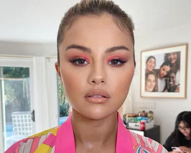 Woman with pink eyeshadow and nude lips in a colorful blouse, standing indoors near a window.