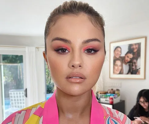 Woman with pink eyeshadow and nude lips in a colorful blouse, standing indoors near a window.