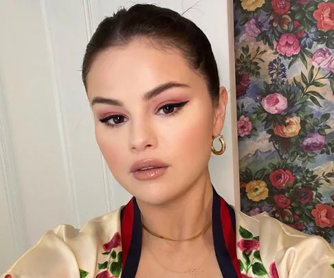 Person with slicked-back hair, wearing gold hoop earrings, in front of a floral patterned background.