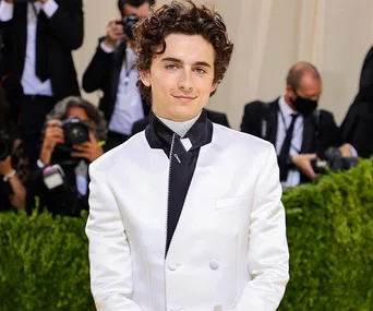 Young man in a white suit smiles amid photographers at an event.