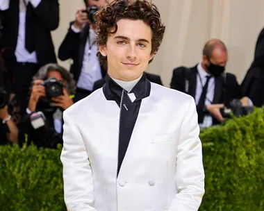 Young man in a white suit smiles amid photographers at an event.