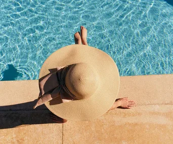 Person in a wide-brimmed hat sitting by a clear blue pool, viewed from above.