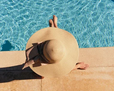 Person in a wide-brimmed hat sitting by a clear blue pool, viewed from above.