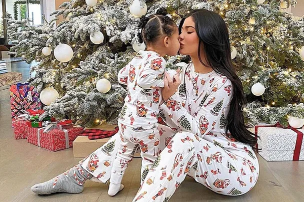 Mother and child in matching Christmas pajamas embrace in front of a decorated tree with gifts underneath.