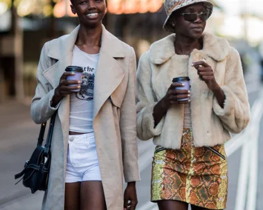 Two fashionable women walking, holding coffee cups; one in a long coat and shorts, the other in a fur jacket and snake-print skirt.