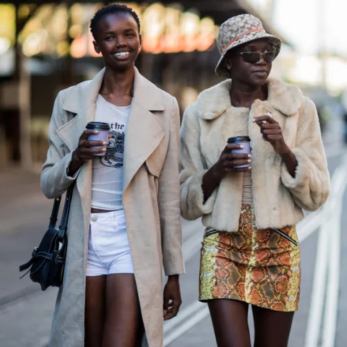 Two fashionable women walking, holding coffee cups; one in a long coat and shorts, the other in a fur jacket and snake-print skirt.