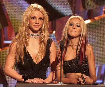 Two women stand on stage with a microphone, dressed in glamorous black outfits, attending an awards event.