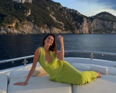 Woman in a bright green dress reclining on a yacht deck against a backdrop of cliffs and ocean.