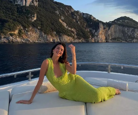 Woman in a bright green dress reclining on a yacht deck against a backdrop of cliffs and ocean.