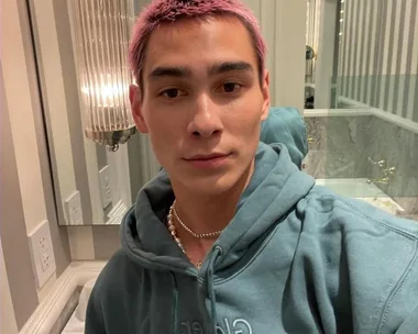 Person with pink hair in a bathroom, wearing a teal hoodie and necklace, facing camera.