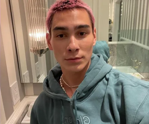 Person with pink hair in a bathroom, wearing a teal hoodie and necklace, facing camera.