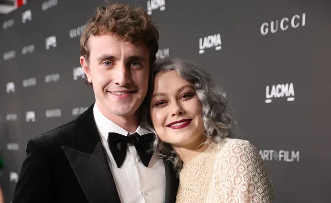 A man in a tuxedo and a woman in a lace dress, smiling together at a formal event.