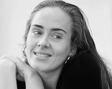 A black and white photo of a smiling woman with long hair, leaning forward with her arms crossed.