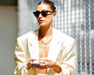 Person in a stylish cream suit, sunglasses, gold jewelry, holding a phone, against a blurred urban backdrop.