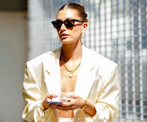 Person in a stylish cream suit, sunglasses, gold jewelry, holding a phone, against a blurred urban backdrop.