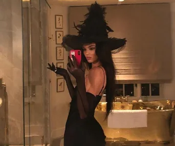 A woman in a black witch costume takes a mirror selfie in a luxurious bathroom.