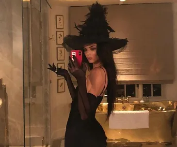 A woman in a black witch costume takes a mirror selfie in a luxurious bathroom.