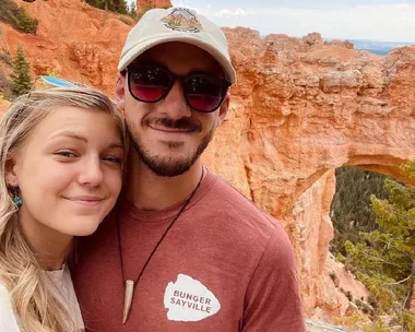 A couple smiling in front of red rock formations and natural arch in a scenic landscape.