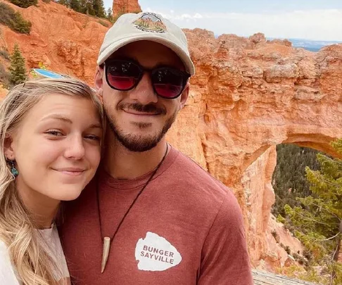 A couple smiling in front of red rock formations and natural arch in a scenic landscape.