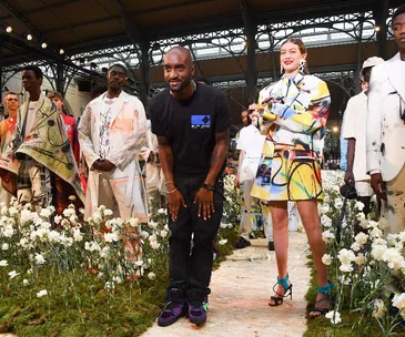 Fashion designer Virgil Abloh bows on runway with models amidst colorful, floral-themed clothing at a fashion show.