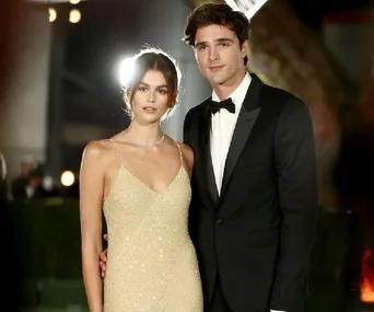 A woman in a shimmering gold gown stands beside a man in a black tuxedo, both posing at a formal event.