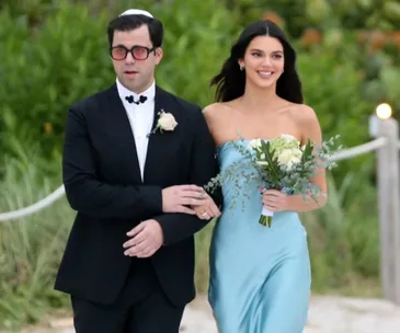 Woman in blue dress holding white flowers walks with man in black suit, outdoors.