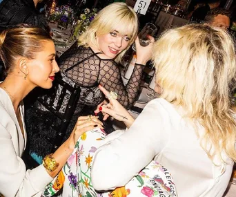 Three women conversing and smiling at an elegant event, decorated table with flowers in the background.