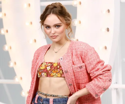 A woman in a pink cardigan and floral crop top poses against a lit background, hands on her hips.