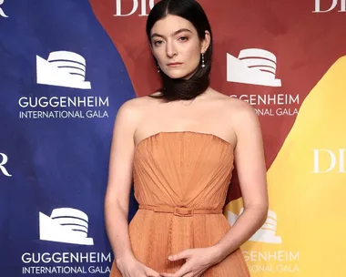 Lorde in an orange gown with hair wrapped around neck, at the Guggenheim International Gala backdrop.