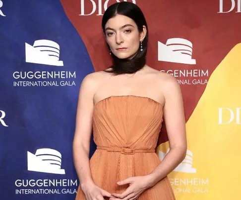 Lorde in an orange gown with hair wrapped around neck, at the Guggenheim International Gala backdrop.