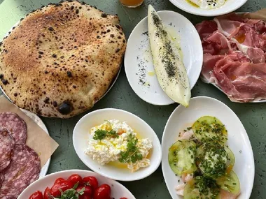 A spread of assorted Italian dishes including pizza, prosciutto, salami, fresh tomatoes, ricotta, burrata, and dressed vegetables.