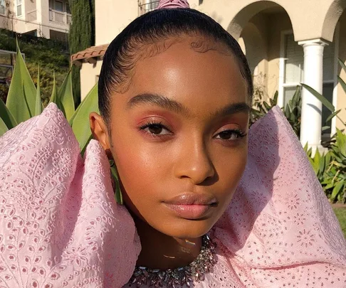 Young woman with slicked hair and pink lace dress stands outdoors, gazing at the camera with a serene expression.