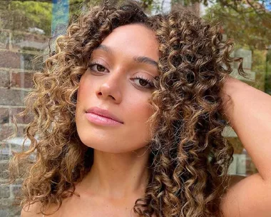 Woman with curly hair touching her head, showcasing natural beauty against a blurred brick background.