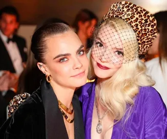 Two women smiling at an event; one with a netted leopard-print hat, the other with slicked-back hair and gold necklace.