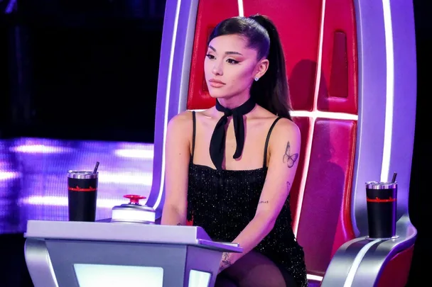 Ariana Grande sits on a red chair in a black glittery dress with a butterfly tattoo on her arm during "The Voice."