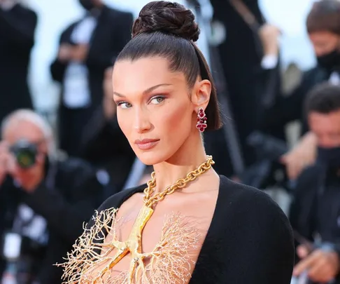 A woman with sleek hair and statement jewelry poses on a red carpet with photographers in the background.