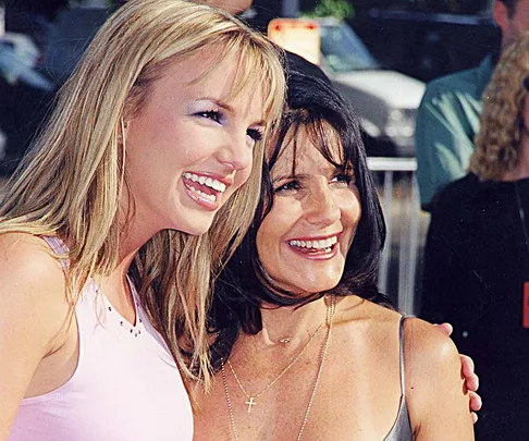 Two women smiling and posing together at an outdoor event, with people blurred in the background.