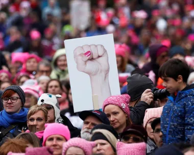 A crowd wearing pink hats, one holding a sign of a fist, during a protest or rally.