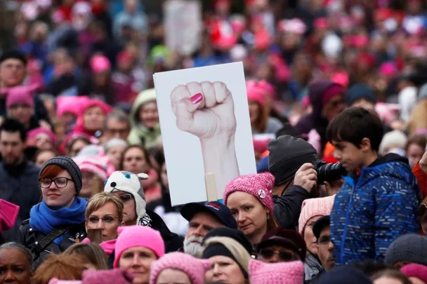 A crowd wearing pink hats, one holding a sign of a fist, during a protest or rally.