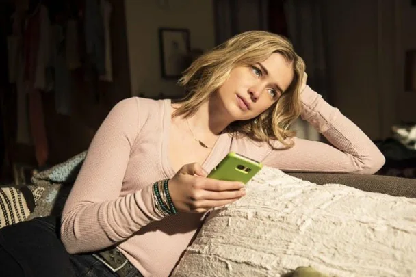 A woman in a pink top holding a green phone, resting on a couch, looking thoughtfully to the side.