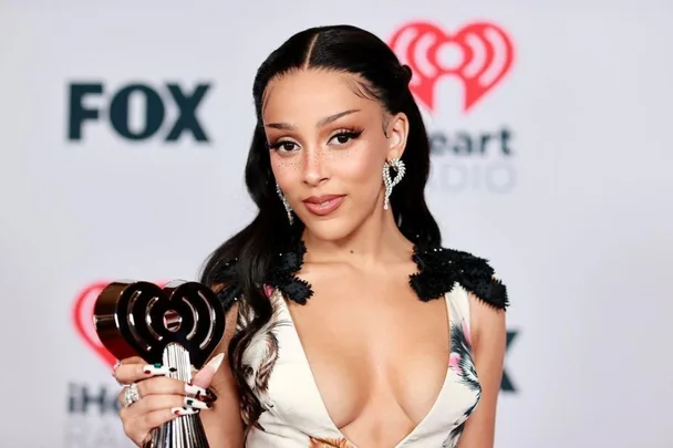 Doja Cat holding an award at the iHeartRadio event, wearing a floral dress with statement earrings.