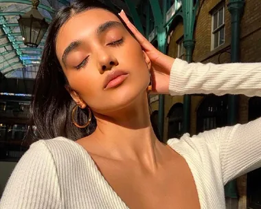 A woman in a white sweater basks in sunlight, eyes closed, with hand on face, standing in an ornate architectural space.