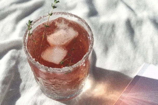 A glass of iced pink drink with a salted rim and herbs, placed on a white cloth in sunlight.