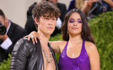 A couple poses at a formal event, she wears a sequined purple dress, he wears a leather jacket without a shirt.